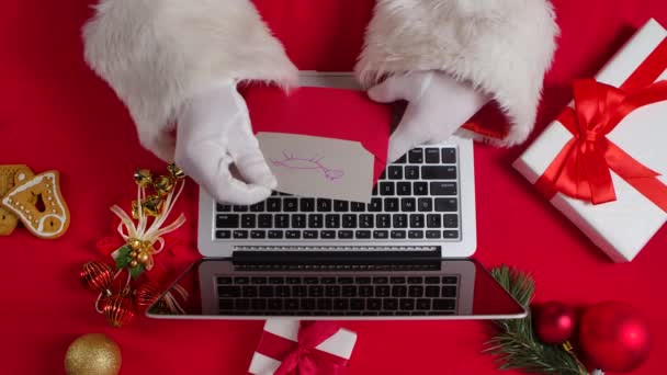 Top view Santa hands in white gloves are typing on keyboard laptop by red New Year decorated table. Santa Claus prohlíží dopisy od dětí a nakupuje dárky na internetu. Zavřít. Zpomalený pohyb. — Stock video