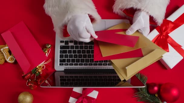 Ovanifrån Santa händer i vita handskar skriver på tangentbordet laptop med rött nyår dekorerat bord. Jultomten tittar igenom brev från barn och köper presenter på Internet. Närbild. Långsamma rörelser. — Stockvideo