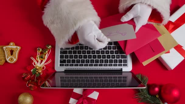 Top view Santa hands in white gloves are typing on keyboard laptop by red New Year decorated table. Santa Claus prohlíží dopisy od dětí a nakupuje dárky na internetu. Zavřít. Zpomalený pohyb. — Stock video