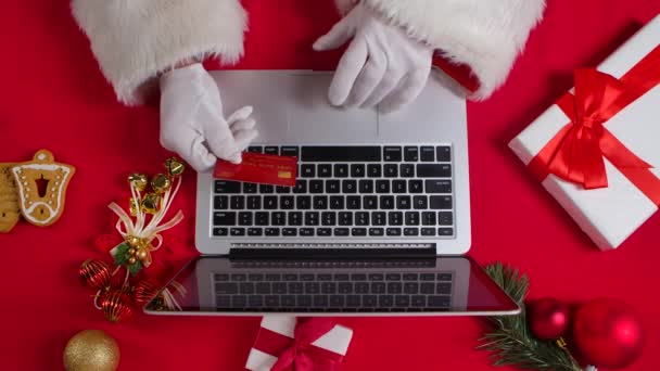 Vista superior Las manos de Santa en guantes blancos están escribiendo en el teclado por la mesa decorada de Año Nuevo rojo. Santa Claus trabaja con un ordenador portátil mientras compra en línea con una tarjeta de crédito. De cerca. Movimiento lento. — Vídeos de Stock