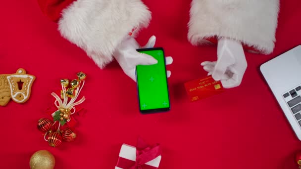 Visão superior Mãos de Papai Noel em luvas brancas usa smartphone com chave de croma de tela verde pela mesa decorada de Ano Novo vermelho. Papai Noel faz compra online com cartão de crédito e telefone. Fecha. Movimento lento. — Vídeo de Stock