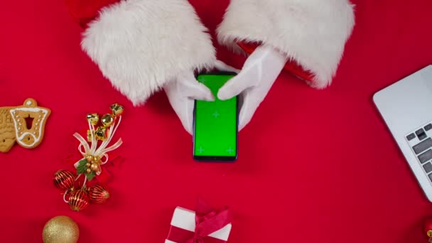 Vue du dessus Les mains du Père Noël utilisent un smartphone avec une clé chromatique à écran vert par une table décorée du Nouvel An rouge. Père Noël tapant sur l'écran pour regarder le contenu, faire des achats en ligne. Ferme là. Mouvement lent. — Video