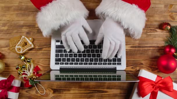 Vista superior Santa manos en guantes blancos escribiendo en el teclado por mesa decorada de madera Año Nuevo. Santa Claus trabaja con el ordenador portátil, mira a través del correo y responde mensajes a los niños. De cerca. Movimiento lento. — Vídeo de stock