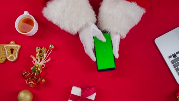 Vista superior Santa manos utiliza teléfono inteligente y bebidas té por mesa roja decorada Año Nuevo. Santa Claus tocando en la pantalla verde croma clave para ver el contenido, hacer compras en línea. De cerca. Movimiento lento. — Vídeos de Stock