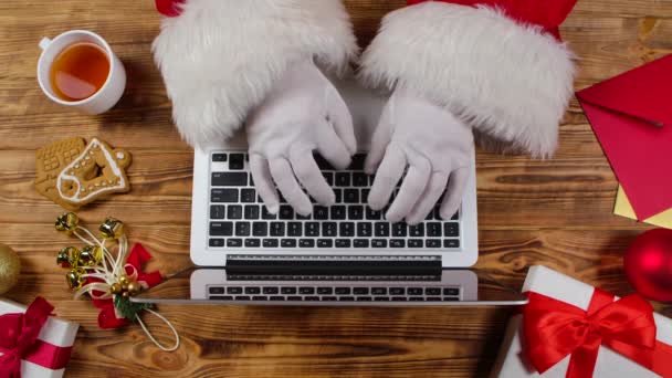 Vue de dessus Santa mains dactylographier sur clavier et boissons thé par table décorée en bois. Père Noël travaille avec ordinateur portable, regarde à travers les lettres des enfants et achète des cadeaux sur Internet. Ferme là. Mouvement lent. — Video