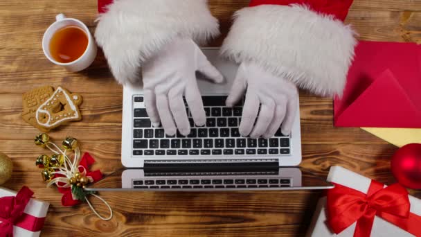 Von oben tippen Weihnachtsmänner auf der Tastatur, trinken Tee an einem mit Holz dekorierten Tisch. Der Weihnachtsmann arbeitet mit Laptop, blättert in Briefen von Kindern und kauft Geschenke im Internet. Aus nächster Nähe. Zeitlupe. — Stockvideo