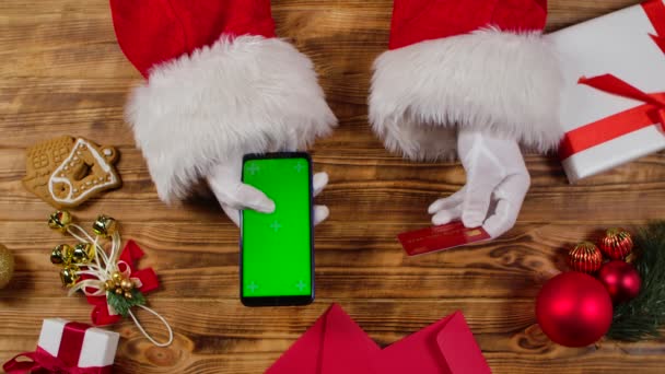 白い手袋のトップビューサンタの手は木製の新年の装飾テーブルで緑の画面クロマキーとスマートフォンを使用しています。サンタクロースはクレジットカードと電話でオンライン購入を行います。閉鎖だ。スローモーション — ストック動画