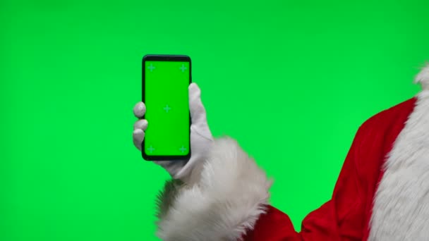 Santa Claus ruce v bílých rukavicích drží smartphone a ukazuje prstem na zelený displej chroma klíč. Izolovaný na zeleném pozadí ve studiu. Zesměšňujte displej. Zavřít. Zpomalený pohyb. — Stock video