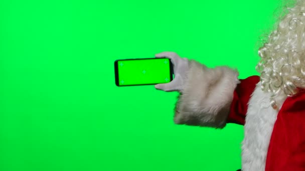 Widok z boku Santa Claus z brodą w białych rękawiczkach trzymać smartfona w pozycji poziomej z zielonym kluczem chroma ekranu. Odizolowany na zielonym tle w studiu. Zamknij drzwi. Zwolniony ruch — Wideo stockowe