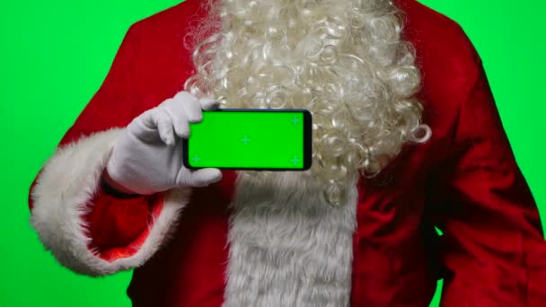 Santa Claus s plnovousem v bílých rukavicích drží smartphone v horizontální poloze a ukazuje prstem zelenou obrazovkou chroma klíč. Izolované na zeleném pozadí ve studiu. Zavřít. Zpomalený pohyb. — Stock video