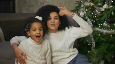 Afro-Amerikan anne ve küçük kızının şok olmuş ve şaşırmış yüz ifadesiyle portresi. Kanepenin yanında oturan kadın ve kız süslü Noel ağacı. Kapatın. Yavaş çekim.