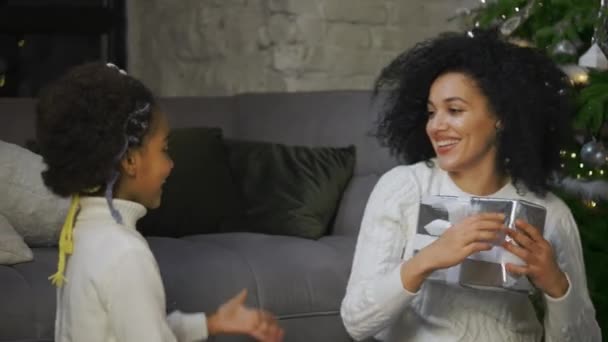 Retrato de mãe afro-americana surpreende sua filhinha com um presente. Mulher e menina sentados perto do sofá e árvore de Natal decorada. Feliz noite de família. Fecha. Movimento lento. — Vídeo de Stock