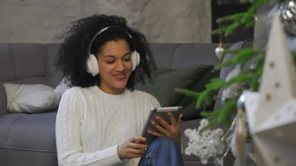 Ritratto di donna afroamericana in grandi cuffie bianche che comunica tramite videochiamata utilizzando un tablet. Giovane donna si siede vicino a un albero di Natale festivo in una stanza luminosa in stile loft. Chiudete. Rallentatore — Video Stock