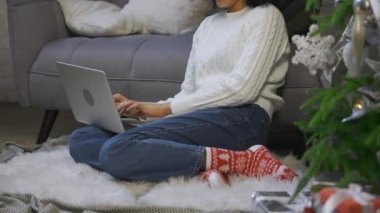 Laptopuyla uzaktan çalışan Afrikalı Amerikalı bir kadının portresi. Genç bir kadın, aydınlık bir çatı katında Noel ağacının yanında oturuyor. Evden işe yarayan kavramsal mesafe. Kapatın. Yavaş çekim