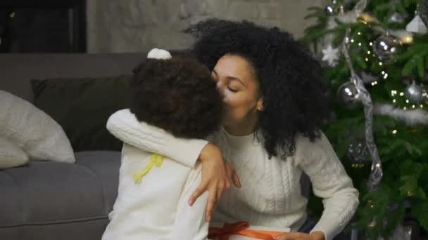 Porträtt av en liten afroamerikansk flicka överraskar sin mor med en gåva. Mor och dotter sitter nära soffan och dekorerade julgran. Trevlig familjekväll. Närbild. Långsamma rörelser. — Stockvideo
