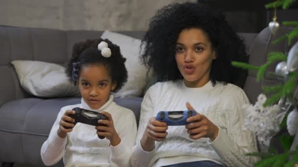 Retrato de mãe afro-americana e sua filhinha jogando o jogo com joysticks e alegria. Mulher e menina sentados perto do sofá e árvore de Natal decorada. Fecha. Movimento lento. — Vídeo de Stock