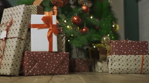 La mujer se acerca al árbol de Navidad y toma la caja de regalo de la pila de regalos que yacen en el suelo de madera. Tarjeta de felicitación navideña. Piernas de mujer en zapatos negros de cerca. Movimiento lento. — Vídeos de Stock