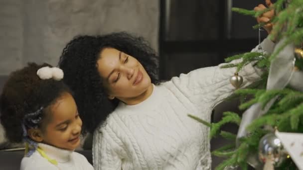 Портрет счастливой афроамериканской мамы и маленькой дочери украшают елку праздничными игрушками, сидя на заднем плане лофт стиле домашней комнаты. Закрывай. Медленное движение. — стоковое видео