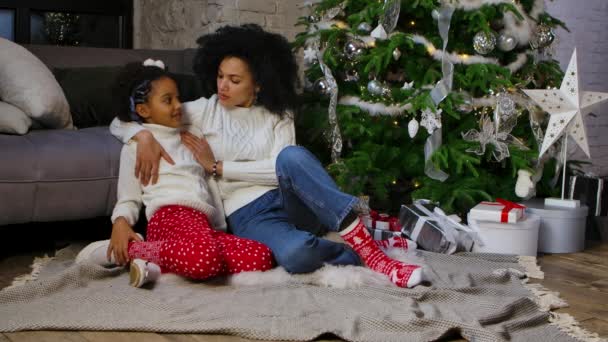 Egy portré afro-amerikai anyáról és a kislányáról, ahogy ölelkeznek és édesen beszélnek. Nő és lány ül közel kanapé és díszített karácsonyfa. Boldog családi estét! Lassú mozgás.. — Stock videók