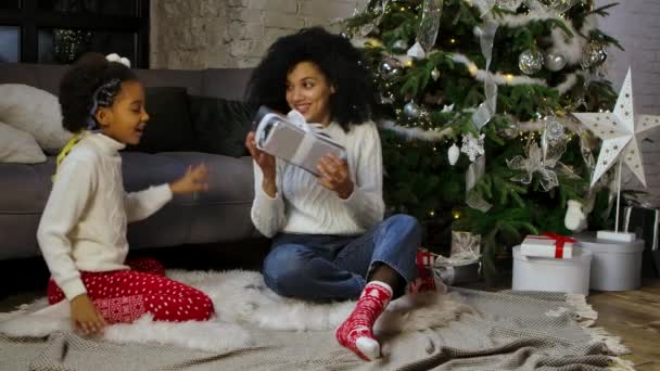 Portret Afroamerykańskiej mamy zaskakuje swoją córeczkę prezentem. Kobieta i dziewczyna siedzi w pobliżu sofy i urządzone choinki. Szczęśliwego rodzinnego wieczoru. Zwolniony ruch. — Wideo stockowe