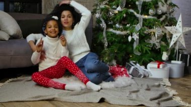 Afro-Amerikan anne ve küçük kızının şok olmuş ve şaşırmış yüz ifadesiyle portresi. Kanepenin yanında oturan kadın ve kız süslü Noel ağacı. Yavaş çekim.