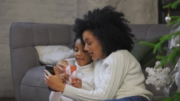 Portret Afroamerykanki i jej córeczki komunikują się przez wideo połączenie za pomocą smartfona. Kobieta i dziewczyna siedzi w pobliżu sofy i urządzone choinki. Zamknij drzwi. Zwolniony ruch. — Wideo stockowe