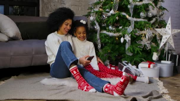 Potret ibu Afrika-Amerika dan putri kecilnya bersenang-senang dan selfie menggunakan smartphone. Wanita dan gadis duduk di dekat sofa dan menghias pohon Natal. Gerakan lambat. — Stok Video
