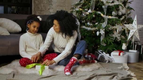 Afrikalı Amerikalı anne ve küçük kızının hediye kutusuna fiyonk bağlarken portresi. Kanepenin yanında oturan kadın ve kız süslü Noel ağacı. Yavaş çekim. — Stok video