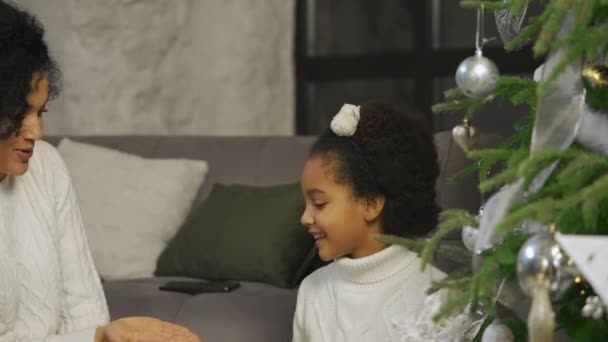Portret szczęśliwej Afroamerykańskiej mamy i córeczki udekorować choinkę świątecznymi zabawkami i bawić się siedząc na tle domu w stylu poddasza. Zamknij drzwi. Zwolniony ruch. — Wideo stockowe
