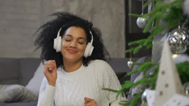 大きな白いヘッドフォンで音楽を聞いて幸せなアフリカ系アメリカ人女性の肖像画。若い女性は明るいロフトスタイルの部屋でお祝いのクリスマスツリーの近くに座っています。お祭り気分。閉めろ。スローモーション — ストック動画