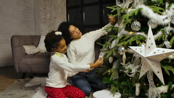 Porträtt av glad afroamerikansk mamma och liten dotter dekorera julgran med festliga leksaker samtidigt sitter mot bakgrund av loft stil hem rum. Trevlig familjekväll. Långsamma rörelser. — Stockvideo