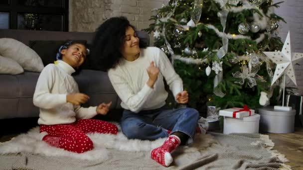 Afrikalı Amerikalı anne ve küçük kızının komik dans ve gülümseme portresi. Kanepenin yanında oturan kadın ve kız süslü Noel ağacı. Mutlu aile akşamları. Yavaş çekim. — Stok video