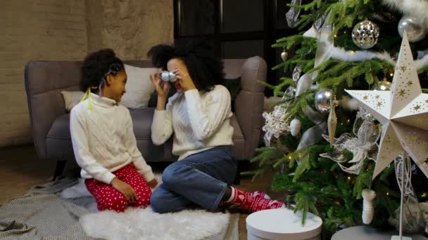 Portret van gelukkige Afro-Amerikaanse moeder en dochtertje versieren kerstboom met feestelijk speelgoed en hebben plezier terwijl ze tegen de achtergrond van loft stijl huiskamer zitten. Langzame beweging. — Stockvideo