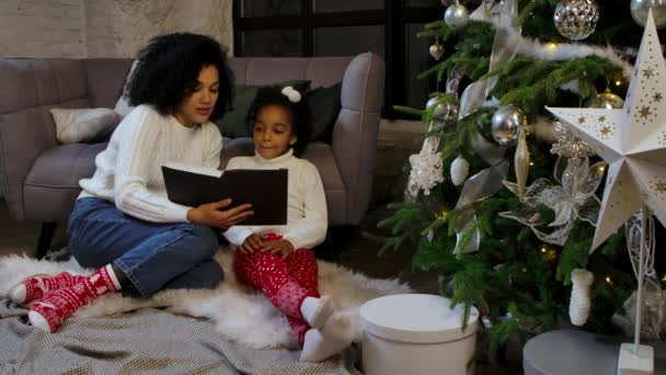 Portrét šťastné afroamerické matky, jak čte pohádku své malé dceři. Žena a dívka sedí u pohovky a zdobené vánoční stromeček. Šťastný rodinný večer. Zpomalený pohyb. — Stock video