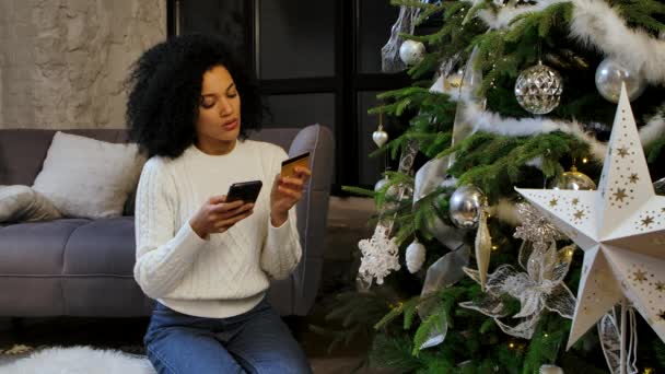 Портрет щасливої афро-американської жінки, яка робить онлайн-покупку, використовуючи свій смартфон і кредитну картку. Молода жінка сидить біля святкового різдвяного дерева у світлому приміщенні. Повільний рух. — стокове відео