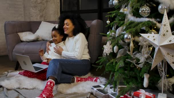 非洲裔美国人妈妈和她的小女儿用笔记本电脑通过视频通话进行交流。坐在沙发旁边装饰圣诞树的女人和女孩。慢动作. — 图库视频影像
