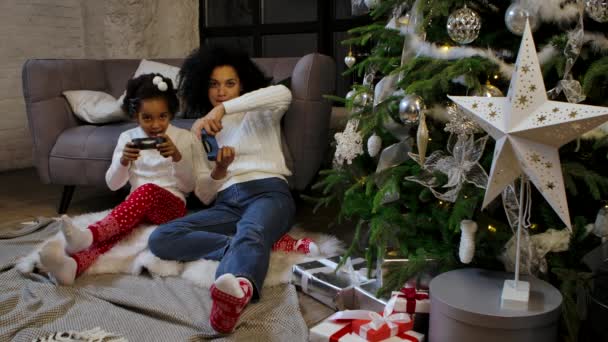 Portrait of African American mom and her little daughter playing the game with joysticks and rejoicing. Woman and girl sitting near sofa and decorated Christmas tree. Slow motion. — Stock Video