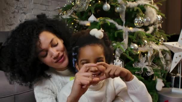 Portrét šťastně se usmívající afroamerické maminky a dcerky, jak se dívají do kamery a dělají prsty gesto srdce. Rodina na pozadí zdobené vánoční stromeček. Zavřít. Zpomalený pohyb. — Stock video