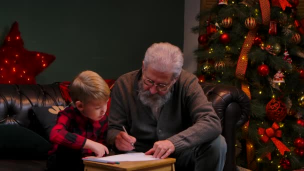 Nagyapa megtanítja az unokáját tollal rajzolni. Egy elegáns ősz hajú férfi egy kisfiúval ül egy kanapén egy díszített szobában, közel egy ragyogó karácsonyfához. Közelről. Lassú mozgás.. — Stock videók