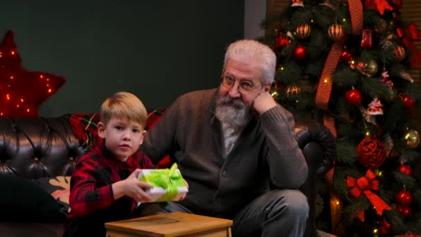 Little boy in a festive red plaid shirt is shake gift box in anticipation of surprise. Grandfather and grandson sit on sofa in a decorated room near glowing Christmas tree. Close up. Slow motion. — Stock Video