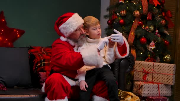 Święty Mikołaj z dzieckiem siedzącym na kolanach komunikują się przez połączenie wideo na smartfonie. Zamknij drzwi. Zwolniony ruch. — Wideo stockowe