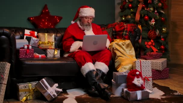 Санта Клаус заказывает подарки в Интернете с помощью ноутбука. Старик с бородой в красном костюме и шляпой сидит на диване в украшенной комнате возле светящейся елки. Медленное движение. — стоковое видео