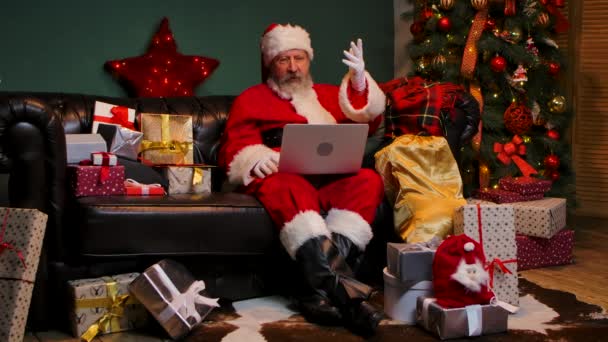 Lindo Santa Claus se comunica por videollamada utilizando un ordenador portátil. Anciano con barba en traje rojo y sombrero se sienta en un sofá en una habitación decorada cerca de un árbol de Navidad brillante y cajas con regalos. Movimiento lento. — Vídeos de Stock