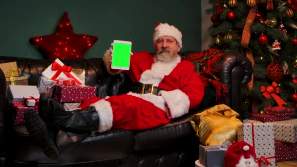 De Kerstman ligt op de bank in de buurt van versierde kerstboom en geschenkdozen, toont tablet met een groen scherm chromen sleutel, waardoor duim omhoog gebaar. Nieuwjaarsreclame, promotie. Langzame beweging. — Stockvideo