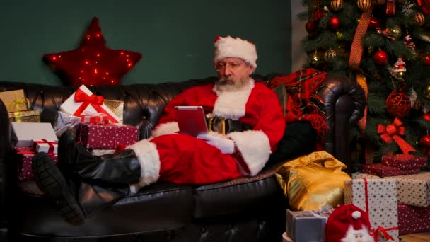 Père Noël communique par appel vidéo à l'aide d'une tablette. Vieil homme avec barbe en costume rouge et chapeau couché sur un canapé dans une pièce décorée près d'un sapin de Noël rougeoyant et des boîtes avec des cadeaux. Mouvement lent. — Video