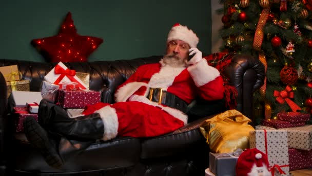 Le Père Noël se trouve sur le canapé près d'un sapin de Noël décoré et des boîtes-cadeaux, parlant sur un smartphone. Félicitations. Concept de l'esprit Nouvel An, fêtes et célébrations. Mouvement lent. — Video