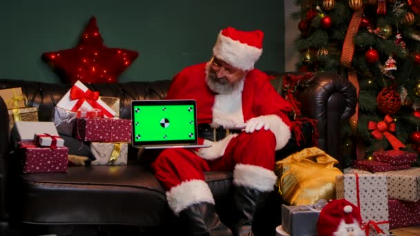 Der Weihnachtsmann sitzt auf dem Sofa neben geschmückten Weihnachtsbäumen und Geschenkschachteln, zeigt Notizbuch mit grünem Chromschlüssel und macht Daumenhoch-Geste. Neujahrswerbung, Promotion. Zeitlupe. — Stockvideo
