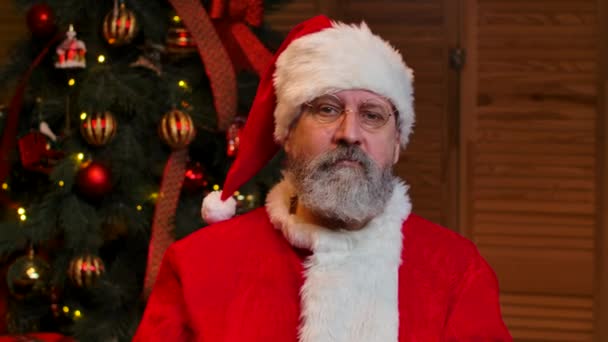 Портрет Санта Клауса серьезно смотрит в камеру, подмигивает и улыбается. Старик с бородой в красном костюме и шляпе на фоне рождественской елки с игрушками и огнями. Закрывай. Медленное движение. — стоковое видео