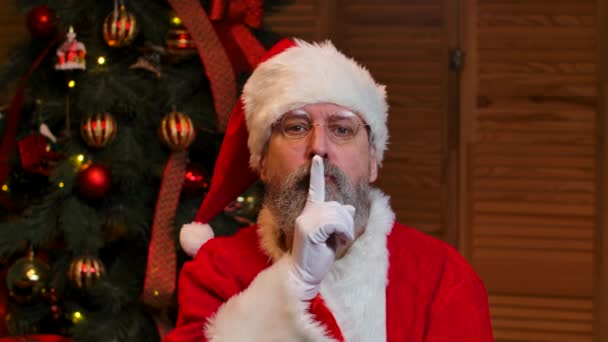 Portret van de Kerstman die gepensioneerd rondkijkt, maakt geheim gebaar en knipoogt. Oude man in rood pak en hoed tegen de achtergrond van kerstboom met speelgoed en verlichting. Sluit maar af. Langzame beweging. — Stockvideo