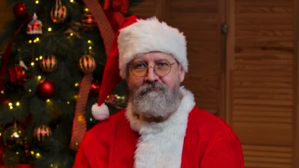 Портрет Деда Мороза, смотрящего в камеру с большим пальцем вверх. Старик с бородой в красном костюме и шляпе на фоне рождественской елки с игрушками и огнями. - Да. Закрывай. Медленное движение — стоковое видео